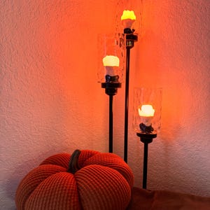 May include: A three-tiered candle holder with three clear glass candle holders and a warm orange glow. The candle holder is on a brown surface with a large orange pumpkin.