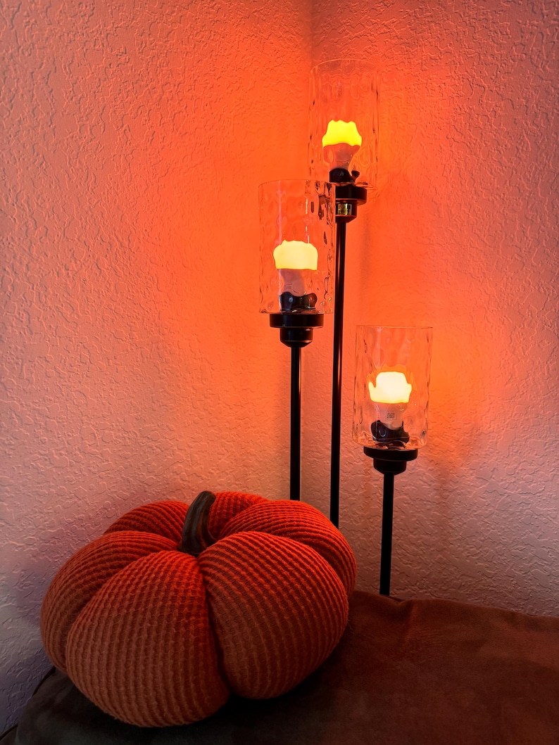 May include: A three-tiered candle holder with three clear glass candle holders and a warm orange glow. The candle holder is on a brown surface with a large orange pumpkin.