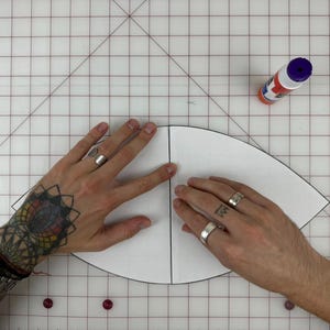 May include: A white paper pattern for a hat is being held by two hands. The pattern is folded in half and the hands are holding the edges of the pattern. A bottle of glue is visible in the upper right corner of the image.