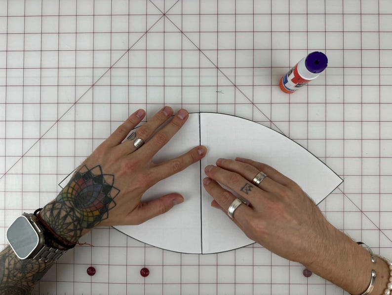 May include: A white paper pattern for a hat is being held by two hands. The pattern is folded in half and the hands are holding the edges of the pattern. A bottle of glue is visible in the upper right corner of the image.