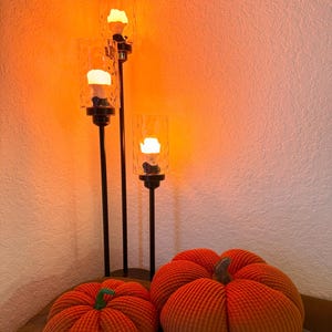 May include: Three glass candle holders with warm orange light bulbs on a black stand with two orange pumpkins in front of them.