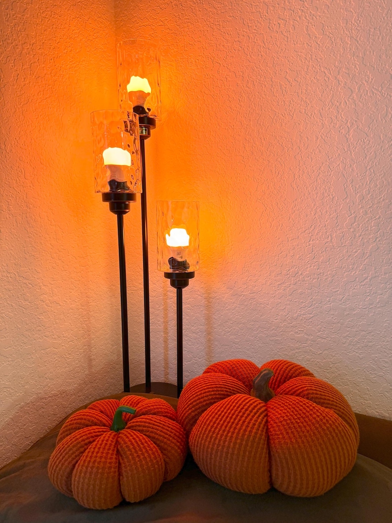 May include: Three glass candle holders with warm orange light bulbs on a black stand with two orange pumpkins in front of them.