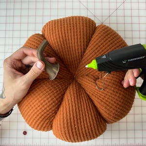May include: A person is attaching a brown metal stem to an orange fabric pumpkin using a hot glue gun. The glue gun is labeled "Surebonder".