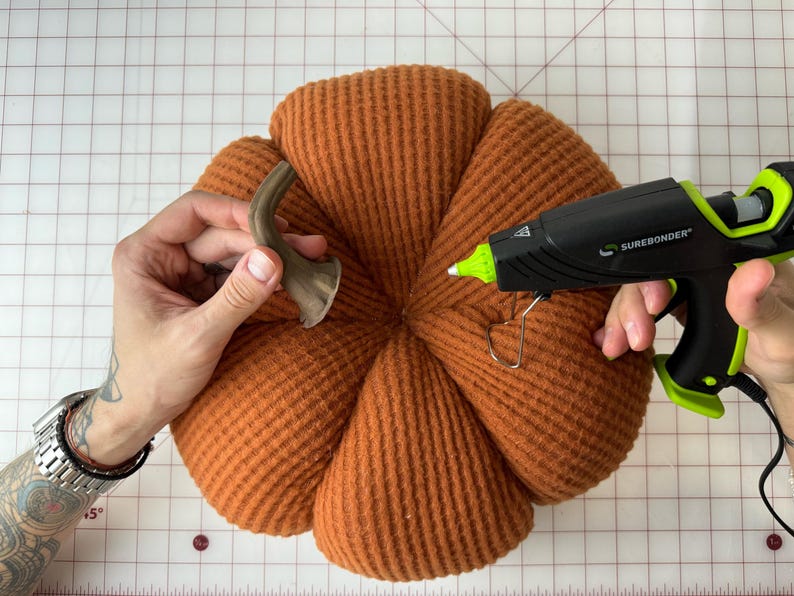 May include: A person is attaching a brown metal stem to an orange fabric pumpkin using a hot glue gun. The glue gun is labeled "Surebonder".