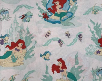 May include: White fabric featuring a repeating pattern of a mermaid with red hair and a green tail, surrounded by sea creatures and aquatic plants. The design includes images of fish, crabs, and a castle, creating an underwater theme.