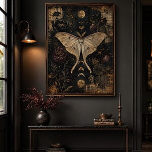 Puede incluir: Una impresi&oacute;n de arte mural rectangular enmarcada que presenta una gran polilla de color crema centrada sobre un fondo oscuro y texturizado. La obra incluye fases lunares doradas, elementos bot&aacute;nicos y texto escrito, todo en un marco de bronce.