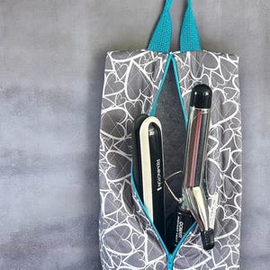 May include: A gray and white heart-patterned travel bag with a teal zipper and handles. Inside the bag are a black and white flat iron and a silver curling iron. The flat iron is labeled "Remington".