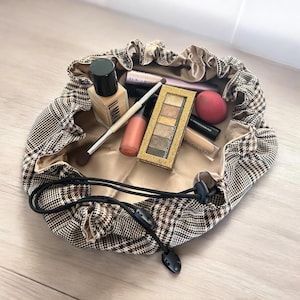 May include: A brown and black plaid makeup bag with a drawstring closure. The bag is open and contains various makeup items, including foundation, lipstick, eyeshadow, and brushes.