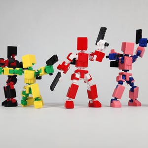 Puede incluir: Cinco figuras de acci&oacute;n articuladas y coloridas posan con armas. Las figuras son rojas, negras, amarillas, rosas y azules. Cada figura tiene una cabeza y un cuerpo en forma de bloque, con articulaciones m&oacute;viles. Est&aacute;n sobre una superficie blanca.