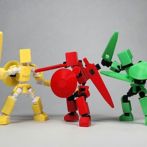 May include: Three toy figures, each with a different color scheme, are posed in a fighting stance. The figure on the left is yellow with a sword and shield. The figure in the center is red with a sword and shield. The figure on the right is green with a sword and shield.