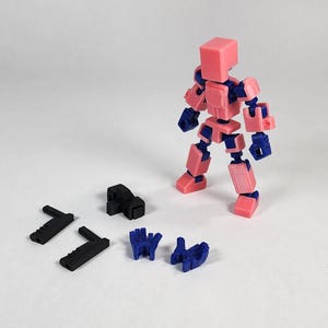 Puede incluir: Una figura articulada rosa y azul con varias piezas negras y azules separadas. La figura est&aacute; de pie, con sus extremidades y cabeza hechas de bloques rosas y articulaciones azules. Hay piezas negras y azules adicionales dispersas cerca.
