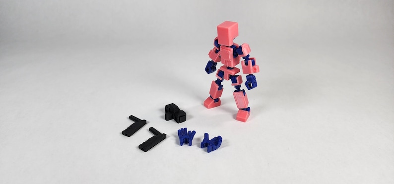 Puede incluir: Una figura articulada rosa y azul con varias piezas negras y azules separadas. La figura est&aacute; de pie, con sus extremidades y cabeza hechas de bloques rosas y articulaciones azules. Hay piezas negras y azules adicionales dispersas cerca.