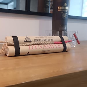 Pode incluir: Um explosivo de dinamite de adereço com tubos de papel bege, faixas pretas e um pavio de corda. Os textos "HIGH EXPLOSIVE" e "DYNAMITE" são impressos nos tubos. Um detonador vermelho está anexado. O adereço está em uma superfície de madeira.