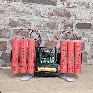 Pode incluir: Um item de brincadeira que se assemelha a uma bomba, com seis bastões de dinamite vermelhos com a inscrição "DYNAMITE". O dispositivo inclui um visor digital que mostra "3.922" e é ligado por fios. Um laptop preto está em primeiro plano.