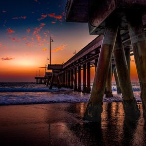 Peut inclure: Une jetée en bois s'étend sur une mer calme au coucher du soleil. Le ciel est d'un orange et d'un rose vifs, avec un croissant de lune visible dans le coin supérieur gauche. La jetée est faite de poutres et de supports en bois robustes, avec quelques lumières visibles le long du sommet.