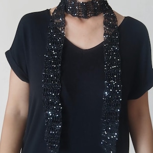 May include: A long, narrow black scarf featuring an open-knit crochet pattern embellished with small, reflective black sequins throughout. The scarf is wrapped around the neck with two long ends hanging down the front over a plain black t-shirt.