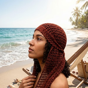 May include: A rust-colored crochet head covering featuring an open, net-like stitch pattern. The headpiece is constructed with a fitted cap section that transitions into two long, dangling side panels that frame the face and shoulders.