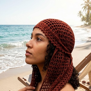 May include: A rust-colored crochet headscarf featuring an open, net-like stitch pattern. The headscarf is designed to cover the top of the head and features long, dangling side panels that drape down, crafted from a textured yarn in a deep earth tone.