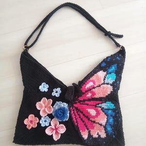 May include: A black crochet handbag with a butterfly design. The butterfly wings are pink, blue, and white. The bag has a black strap and is decorated with small crochet flowers in pink and blue.