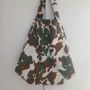 Könnte beinhalten: Eine Tragetasche mit Camouflage-Muster in den Farben Waldgrün, Braun und Off-White. Die Tasche hat eine breite, dreieckige Form mit zwei integrierten Schulterriemen an der Oberseite und besteht aus einem strapazierfähigen Stoff mit sichtbarem Gewebe.