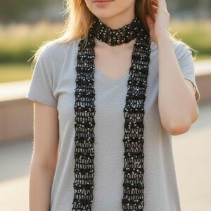 May include: A long, narrow black scarf featuring an open-knit crochet pattern embellished with small, reflective silver sequins throughout. The scarf is wrapped around the neck and hangs down in two parallel vertical strips over a grey V-neck t-shirt.