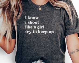 May include: Dark gray t-shirt with the text "i know i shoot like a girl try to keep up" in white. The shirt has short sleeves and a crew neck. A gold necklace is visible.