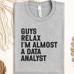 May include: A heather gray crew neck t-shirt with black text printed on the front that reads: GUYS RELAX I'M ALMOST A DATA ANALYST. The shirt features a standard short-sleeve design with a white brand label visible inside the collar area.