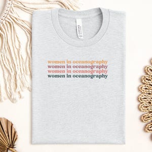 Può includere: Una maglietta grigia chiaro con il testo "women in oceanography" ripetuto quattro volte in diverse tonalità di rosa, arancione e grigio.