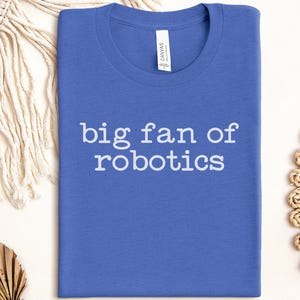 May include: A blue t-shirt with the text "big fan of robotics" in white. The shirt is folded and centered in the image. The shirt has a crew neck and a tag that says "CANVAS".