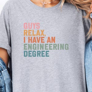 May include: A heather gray t-shirt with the text "GUYS RELAX, I HAVE AN ENGINEERING DEGREE" in pastel colors. The shirt has rolled-up sleeves and is worn with a denim jacket.