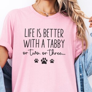 May include: A light pink t-shirt with the text "LIFE IS BETTER WITH A TABBY or two, or three..." in black. Below the text are three black paw prints. The shirt is being worn with a denim jacket.