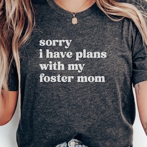 May include: Dark gray t-shirt with the text "sorry i have plans with my foster mom" in white. The shirt is a crew neck and has short sleeves. A gold necklace is visible.