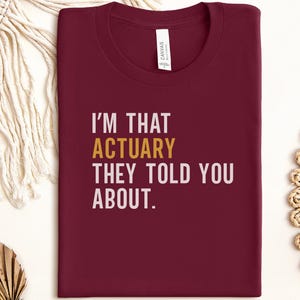 May include: A maroon t-shirt with the text "I'M THAT ACTUARY THEY TOLD YOU ABOUT." The word "ACTUARY" is in gold, and the rest of the text is white. The shirt is folded neatly.
