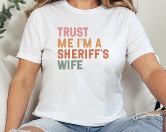 May include: White t-shirt with the text "TRUST ME I'M A SHERIFF'S WIFE" in pink, orange, and green block letters. The shirt is short-sleeved and appears to be made of cotton.