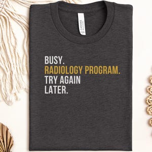 May include: A dark charcoal gray crew neck t-shirt featuring the white and gold text BUSY. RADIOLOGY PROGRAM. TRY AGAIN LATER. The shirt has a standard fit with a visible brand label inside the collar area, showcasing a simple graphic design.