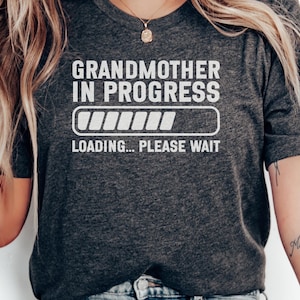 May include: A dark heather gray short-sleeve t-shirt featuring white text that reads GRANDMOTHER IN PROGRESS with a loading bar graphic below it and the additional text LOADING... PLEASE WAIT centered on the front of the garment.