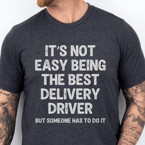 May include: A dark heather grey short-sleeved t-shirt featuring the white distressed text: IT'S NOT EASY BEING THE BEST DELIVERY DRIVER BUT SOMEONE HAS TO DO IT. The shirt has a crew neckline and a standard fit, displaying the printed message clearly.
