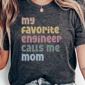 Puede incluir: Camiseta gris oscuro con el texto "my favorite engineer calls me mom" en colores pastel. La camiseta es de cuello redondo y tiene mangas cortas. El texto está apilado verticalmente.