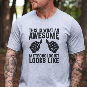 May include: A light gray t-shirt with the text "THIS IS WHAT AN AWESOME METEOROLOGIST LOOKS LIKE" in black. The design includes two thumbs-up gestures. The shirt is made of a soft material.