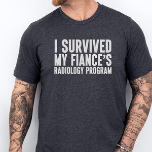 May include: A dark gray t-shirt with the text "I survived my fiance's radiology program" printed in white.