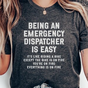 May include: Dark gray t-shirt with the text "BEING AN EMERGENCY DISPATCHER IS EASY." The shirt also has the text "IT'S LIKE RIDING A BIKE EXCEPT THE BIKE IS ON FIRE YOU'RE ON FIRE EVERYTHING IS ON FIRE."