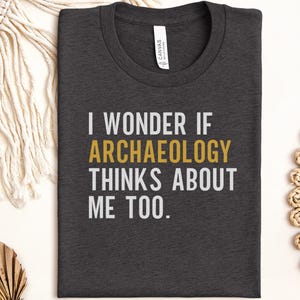 Peut inclure: Un t-shirt à col rond gris anthracite foncé avec le texte blanc "I WONDER IF", suivi du texte doré "ARCHAEOLOGY" et du texte blanc "THINKS ABOUT ME TOO.". Une étiquette de marque blanche est visible à l'intérieur du col.