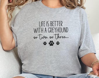May include: A light gray t-shirt with the text "LIFE IS BETTER WITH A GREYHOUND or Two, or Three..." in black script. Below the text are three black paw prints. The shirt is a casual style.