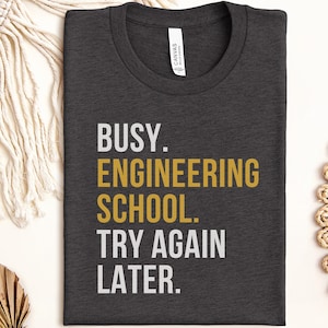 May include: A dark charcoal grey crew neck t-shirt featuring the white and gold text BUSY. ENGINEERING SCHOOL. TRY AGAIN LATER. The shirt has a white brand label at the inner neckline and is made of a soft, heathered fabric with a simple design.