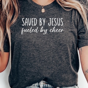 May include: Dark gray t-shirt with the text "SAVED BY JESUS fueled by cheer" in white. The shirt has a crew neck and short sleeves. The image shows the upper body of a person wearing the shirt.