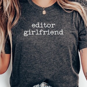 May include: Dark gray t-shirt with the words "editor girlfriend" printed in white. The shirt is a crew neck style and has short sleeves. The image shows the upper body of a person wearing the shirt.