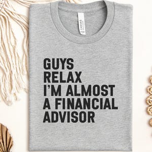May include: A heather gray crew neck t-shirt featuring the black printed text GUYS RELAX I'M ALMOST A FINANCIAL ADVISOR. The shirt has a standard short-sleeve design with a visible brand label inside the collar area on the inner neckline.