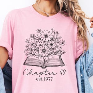 May include: A light pink t-shirt featuring a black and white floral design atop an open book. The text "Chapter 49 est. 1977" is written below the book in a script font. The shirt has short sleeves.