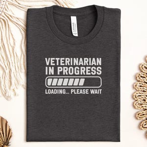 Puede incluir: Camiseta gris oscuro con el texto blanco "VETERINARIAN IN PROGRESS" y una barra de carga. Debajo, el texto dice "LOADING... PLEASE WAIT". La camiseta doblada descansa sobre una superficie neutra.