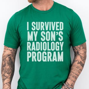 May include: A green t-shirt with the text "I SURVIVED MY SON'S RADIOLOGY PROGRAM" in white block letters. The shirt is a crew neck style and is being worn by a person with tattoos on their arms.
